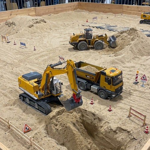 Detailed shot of a yellow RC excavator moving dirt next to an RC dump truck on a miniature construction site, realistic details, focused lighting, shallow depth of field.